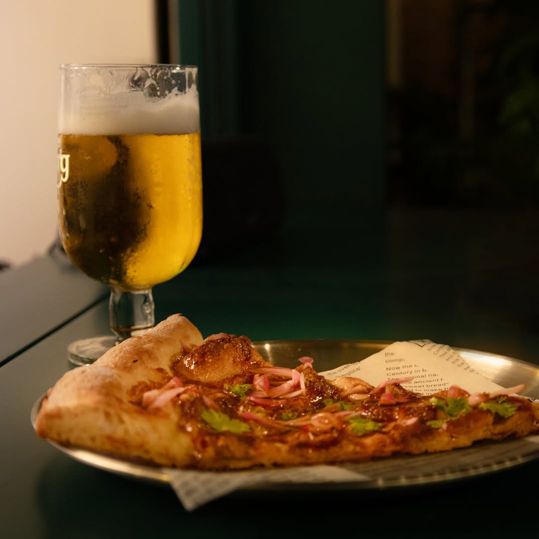 A folded slice of New York style pizza on a metal tray beside a cold beer.