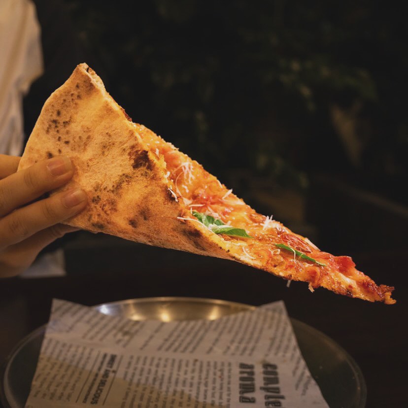 A hand holding a folded slice of New York style pizza, blistered crust, basil and parmesan on top.
